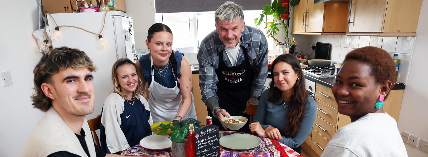 Six people in a shared student kitchen sitting at a table looking to the camera and smiling, about to eat a meal together.
