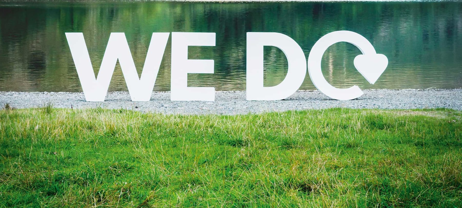 Large white letters on the shore of a lake spelling out WE DO with the O replaced with the recycling swoosh