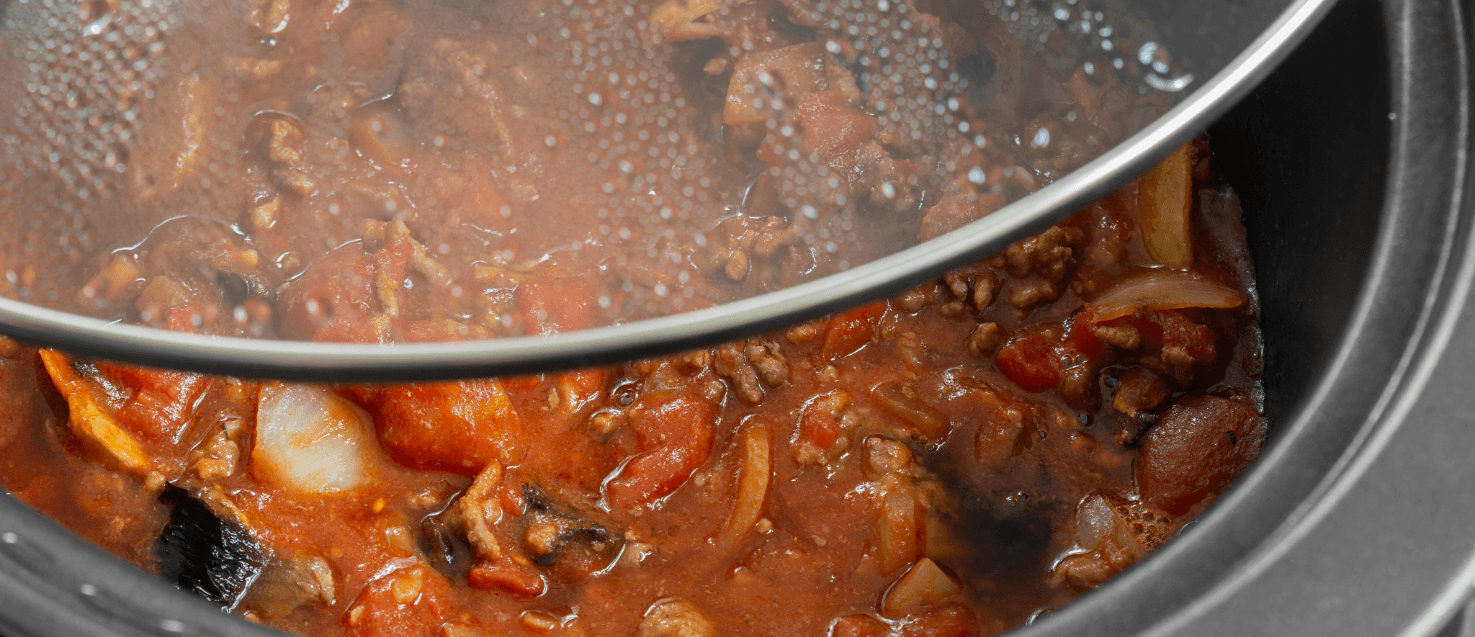 close-up of a slow cooker with tasty mince ina tomato sauce withonions and over veggies and herbs